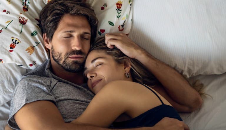 A couple holds each other in bed as they sleep. The pillow behind them features a floral pattern and the person on the left is smiling with their eyes shut. This photo is being used in an article about sexual personality types.