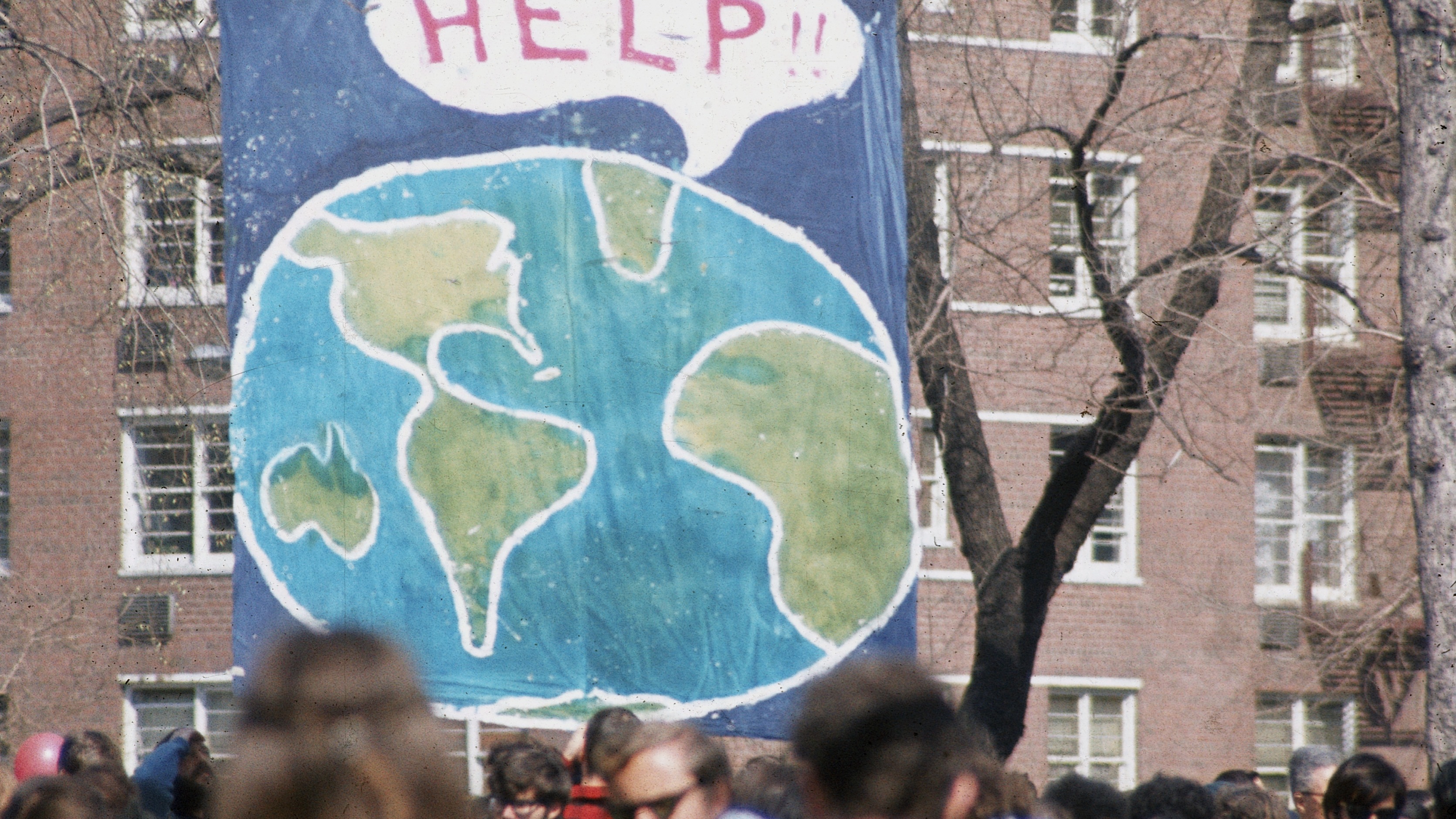 A crowd of people gather near a large poster that shows a speach bubble from planet Earth that reads 'Help!!', on the occaision of the first Earth Day conservation awareness celebration, New York, New York, April 22, 1970.