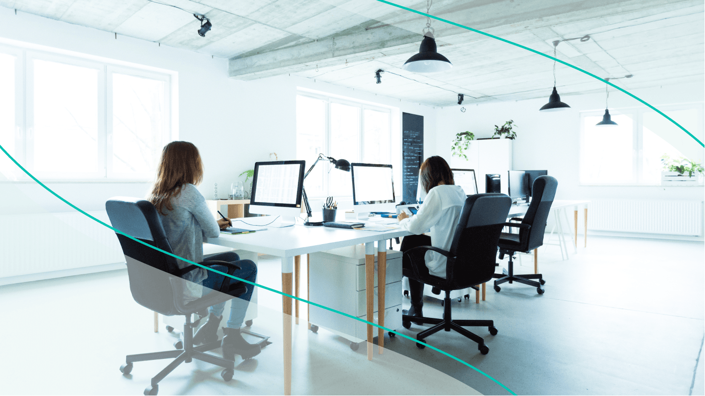 Women work at shared desk cluster, their backs to the camera.