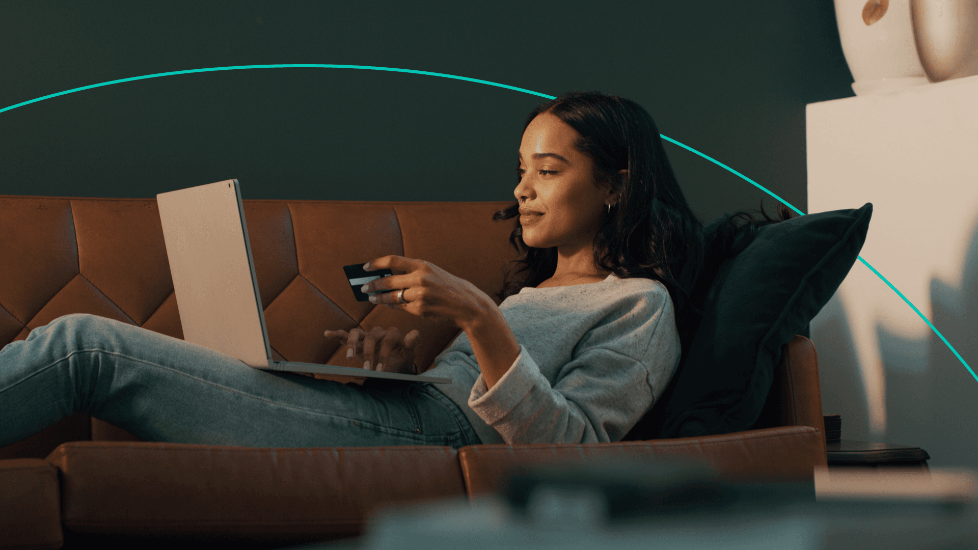 A woman lies on her couch with a laptop in her lap and a credit card in one hand while the other hand is typing.