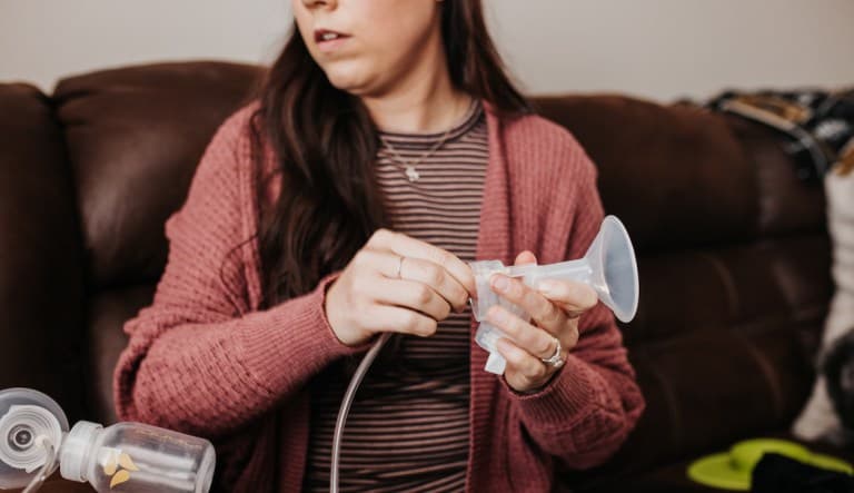 A mom sits on the couch holding a breast pump and preparing to pump. She is looking off to the side as if to make sure she has all the tools she needs. This photo is being used in an article about sad nipple syndrome.