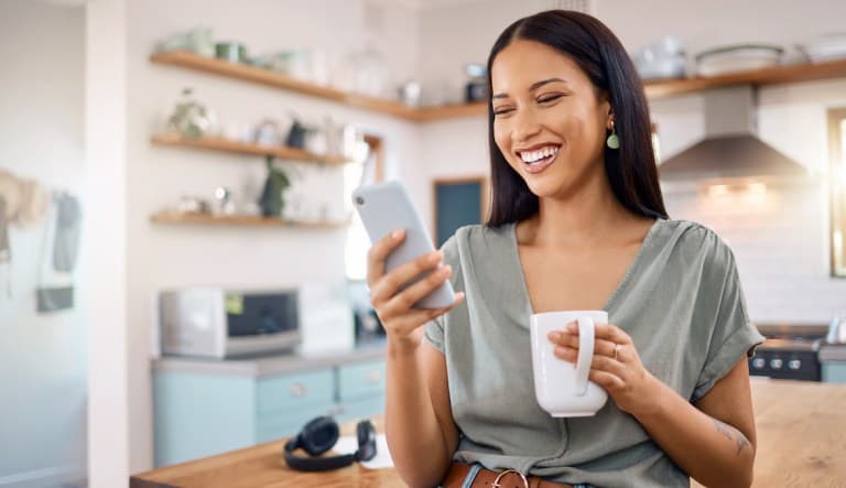 A woman stands in her kitchen, smiling at her phone and enjoying a cup of coffee. This photo is being used in an article about dating profiles.