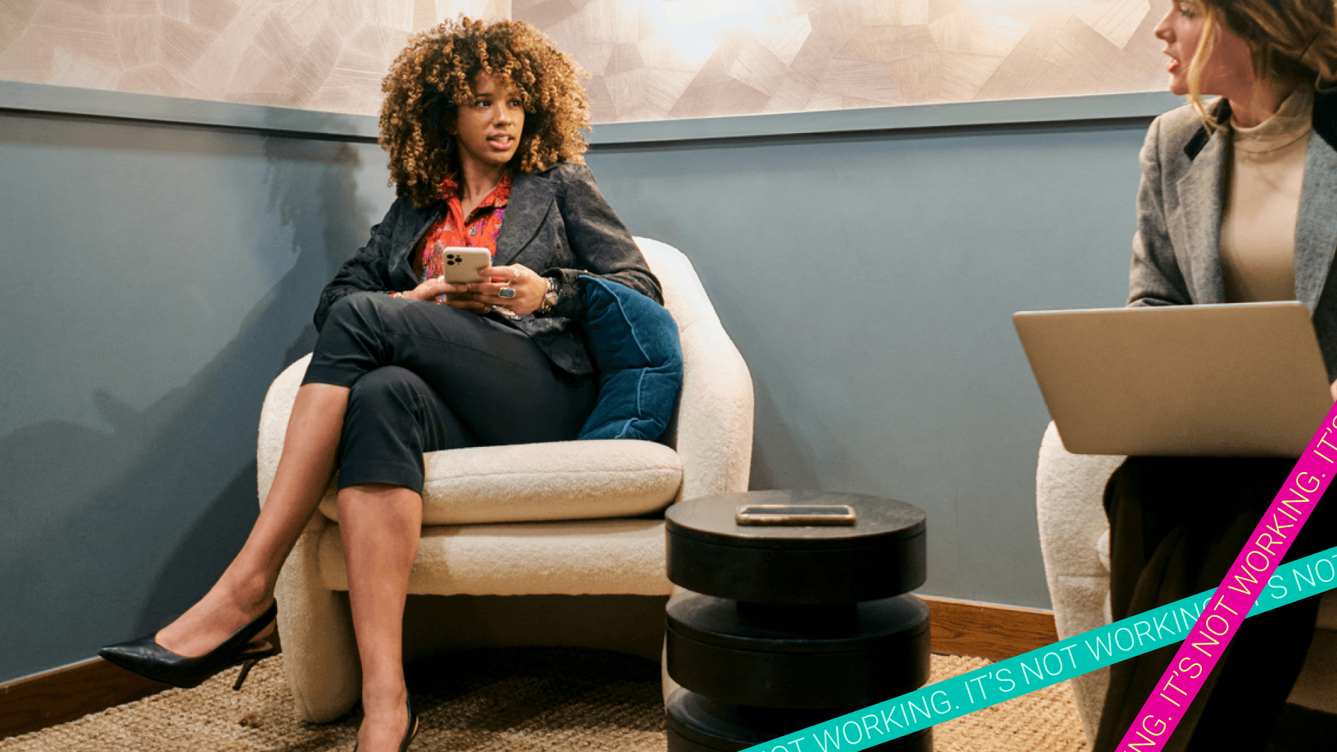 Woman in work attire holds her phone while chatting with another woman using a laptop. Women say they are increasingly shaping their careers to benefit their wellbeing.