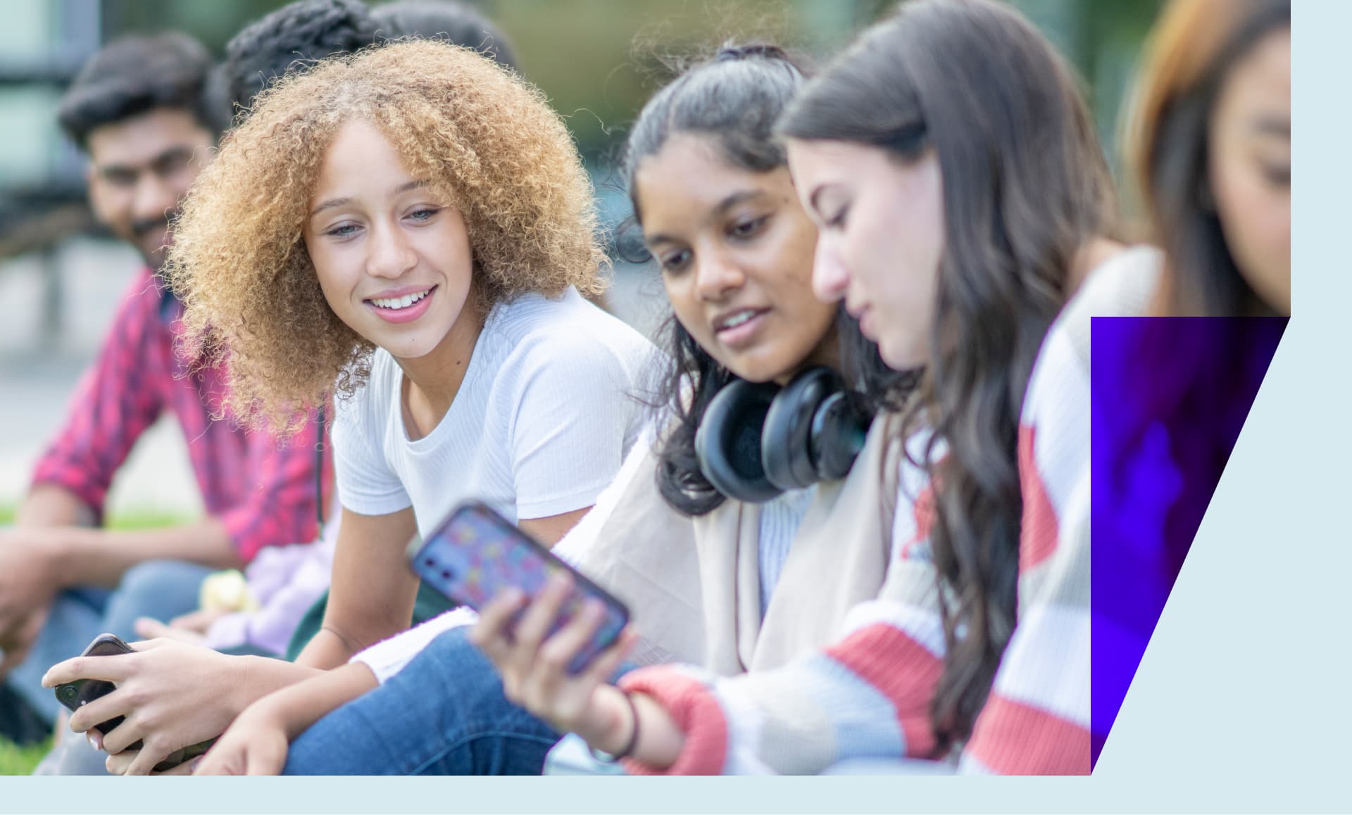 A group if University students sit on the grass outside on campus as they take a break between classes. They are each dressed casually and have their cell phones out as they catch up on social media.