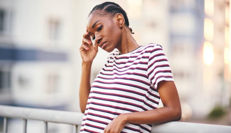 A Black woman in a striped shirt leans against railing and touches one hand to her temple as if in distress. This photo is being used to promote an article answering the question