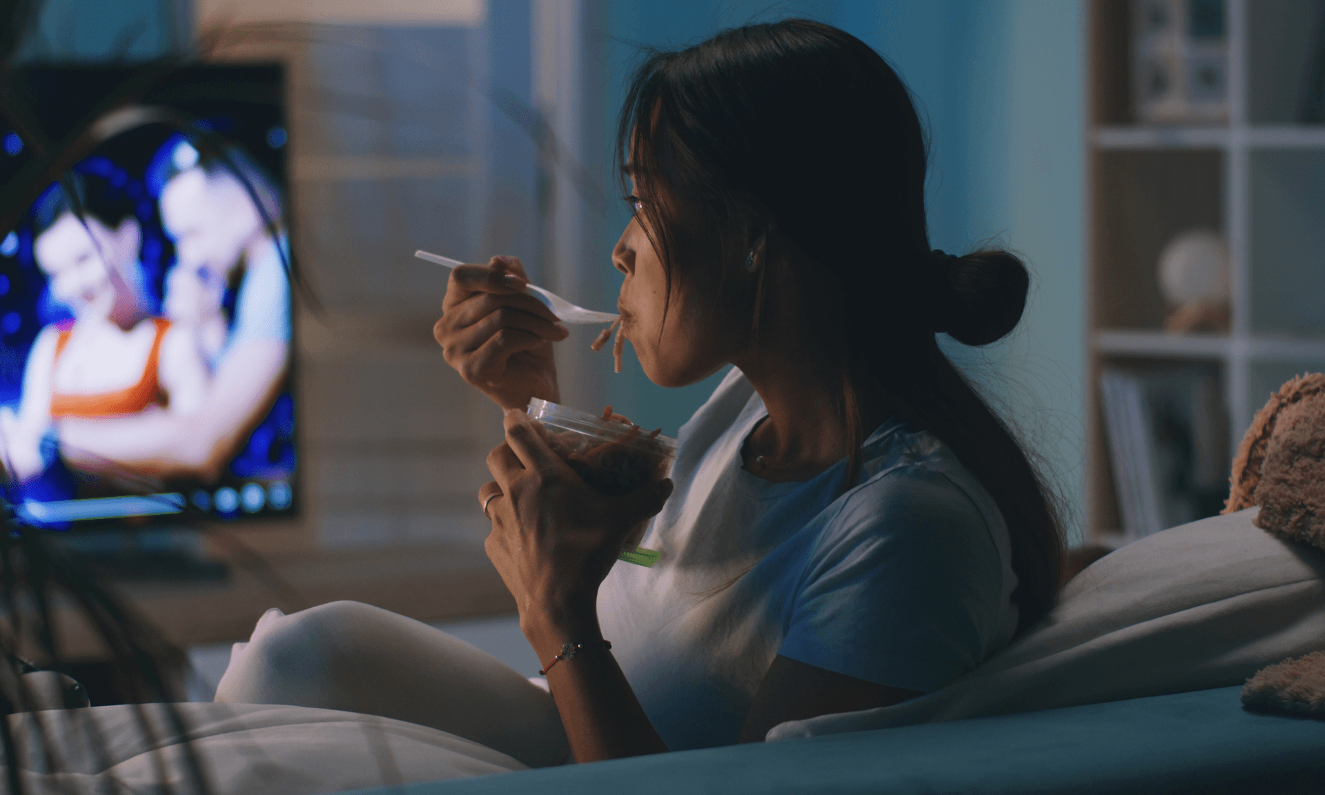 A woman sitting on her couch eating noodles and watching TV
