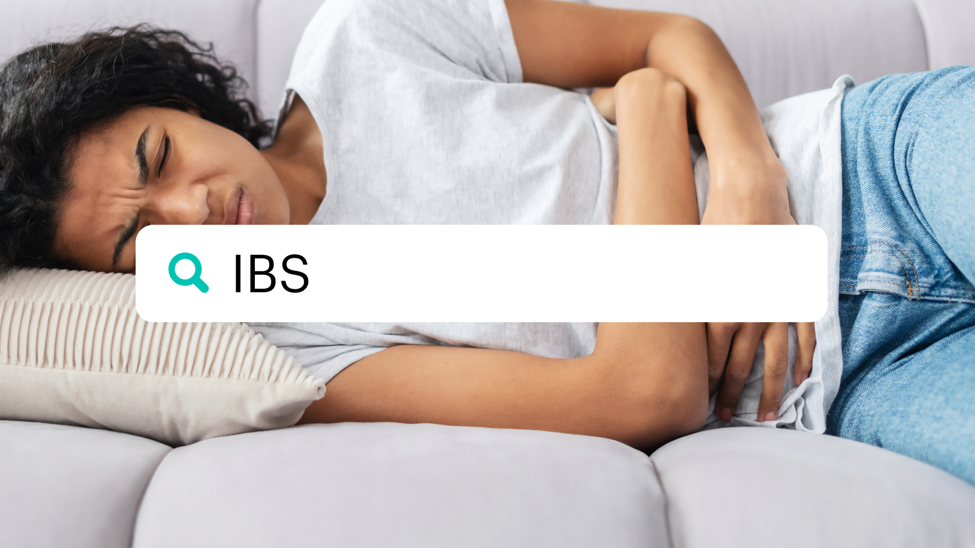 Woman lies on the couch while clutching her stomach, in front of a search bar that says "IBS"