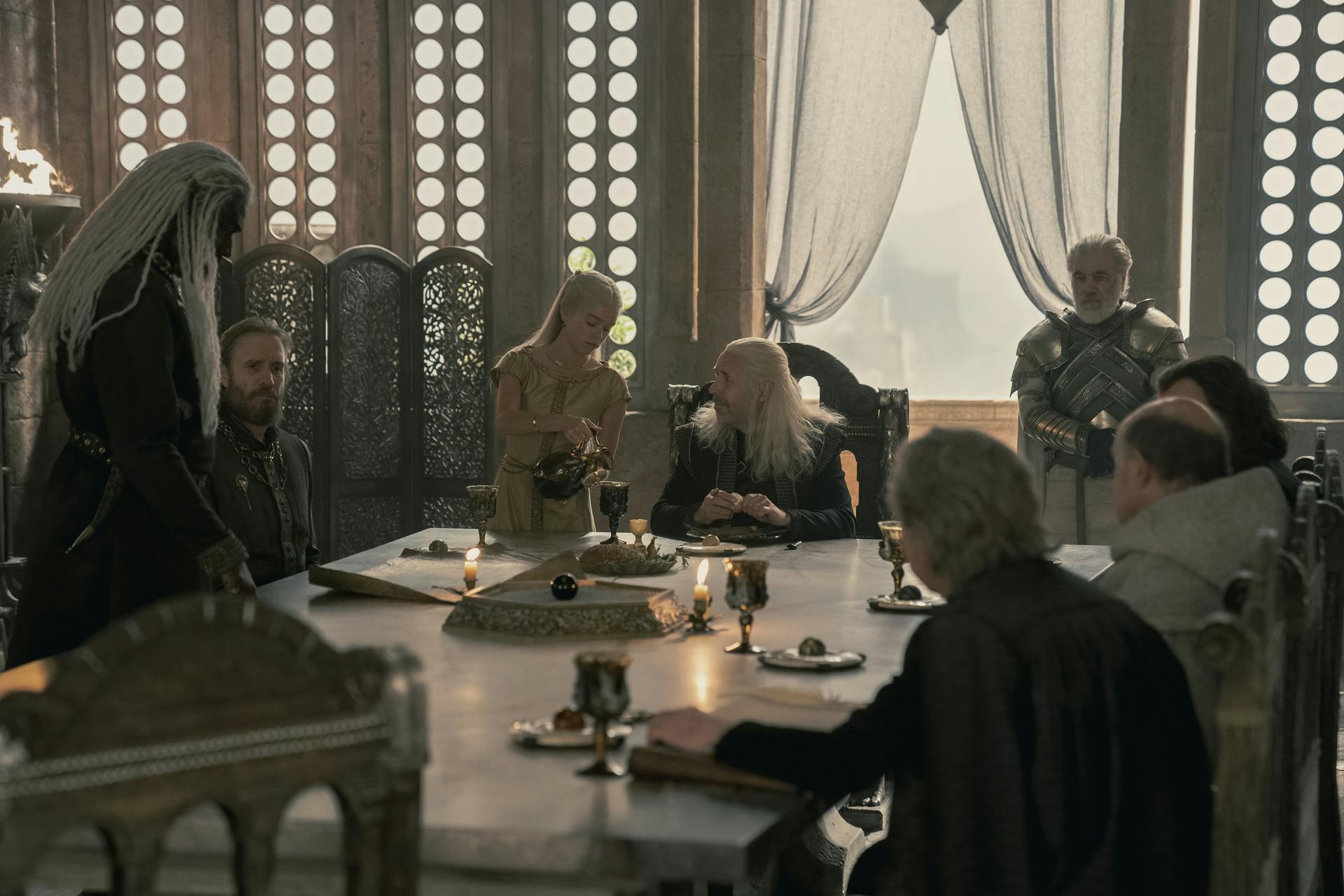 King Viserys Targaryen at a table as Princess Rhaenyra pours him a drink.