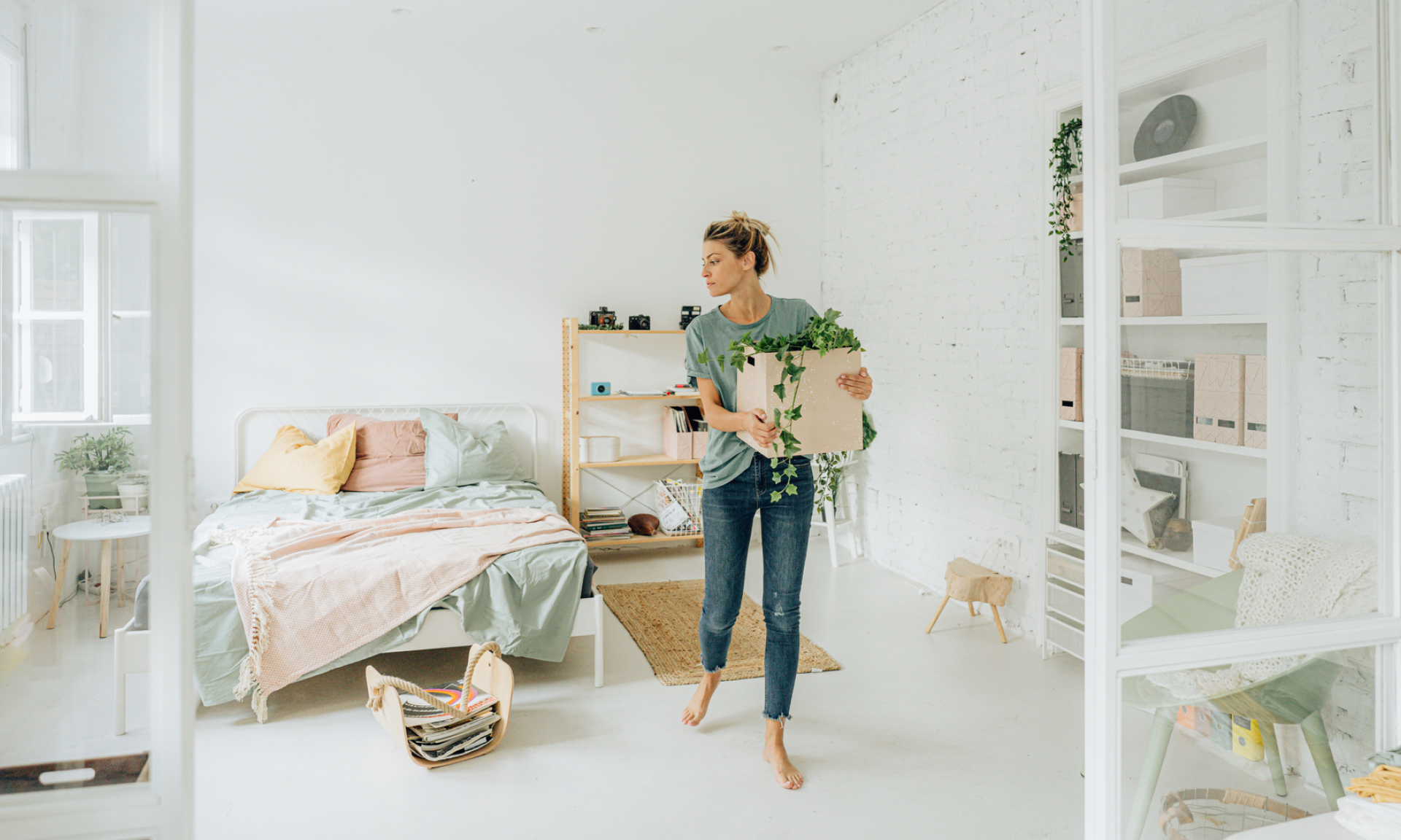 A woman carrying a box with a plant in it out of a very clean, white bedroom